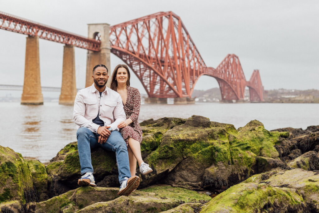 A couple sit with the Forth Rail Bridge behind them during their Edinburgh engagement photos.